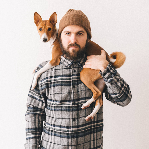Young white bearded man in a grey plaid shirt and knit hat holding his ginger african basenji dog friend. Friendship between animals and people. - Photo, Image