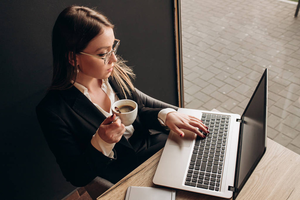 una hermosa chica vestida con ropa de oficina con gafas de sol sentada y sosteniendo una taza de café y escribiendo en el portátil en la cafetería
 - Foto, Imagen
