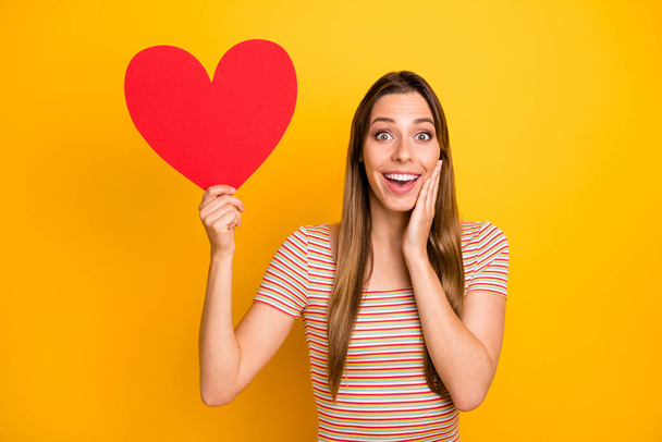 Photo of funny attractive lady hold big red paper heart date invitation not believe happiness arm on cheek wear casual striped t-shirt isolated yellow color background - Foto, immagini