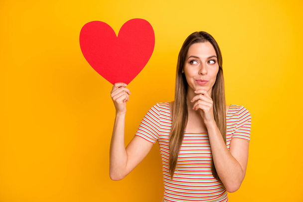 Photo of attractive pretty lady hold big red paper heart date invitation think go or not cunning look side arm on chin wear casual striped t-shirt isolated yellow color background - Foto, Imagen