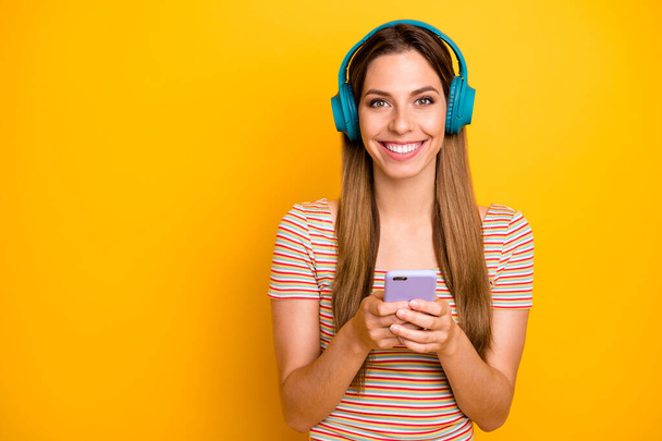 Photo of beautiful lady holding telephone listen music in cool wireless earphones choosing next song wear casual striped t-shirt isolated yellow color background - Foto, Imagem