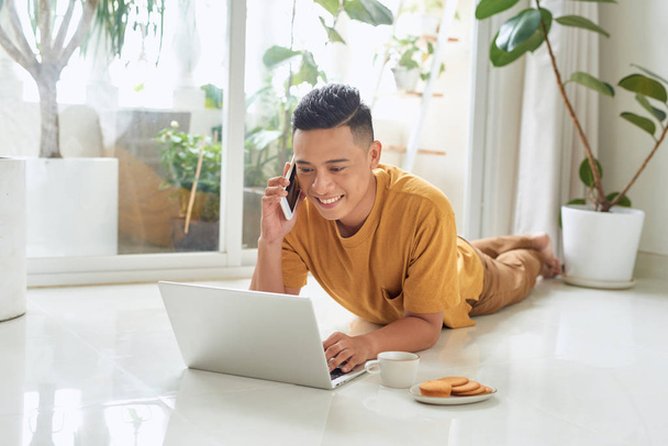 Усміхнений чоловік розмовляє на мобільному телефоні і використовує ноутбук, лежачи на підлозі вдома
 - Фото, зображення