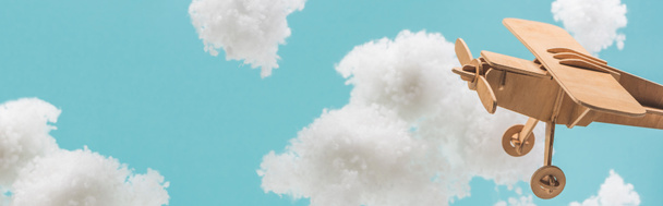 青いパノラマのショットに隔離された綿ウールで作られた白いふわふわの雲の間を飛ぶ木製のおもちゃの飛行機 - 写真・画像