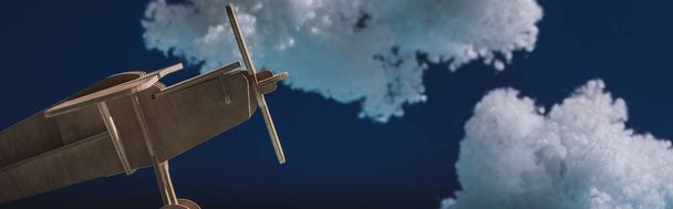 drewniany samolot zabawkowy latający wśród białych puszystych chmur wykonanych z wełny bawełnianej izolowanej na ciemnoniebieskim, panoramicznym ujęciu - Zdjęcie, obraz