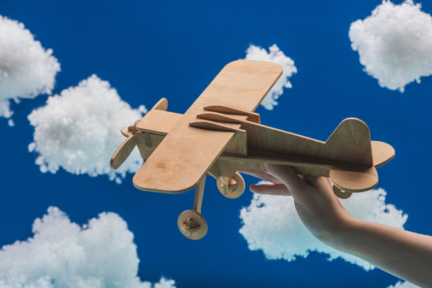 青い上に隔離された綿ウールで作られた白いふわふわの雲の近くに木製のおもちゃの飛行機を保持する女性のクロップドビュー - 写真・画像