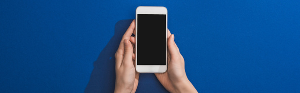 cropped view of woman holding smartphone on blue background, panoramic shot - Zdjęcie, obraz