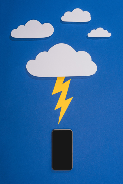 top view of white paper cut clouds with lighting hitting smartphone on blue background - Foto, imagen