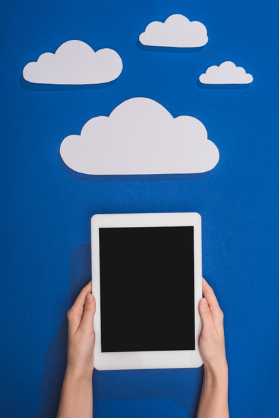 cropped view of woman holding digital tablet near white paper cut clouds on blue background - Valokuva, kuva