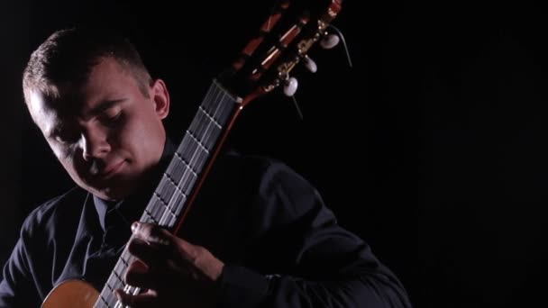 όμορφος νεαρός παίζει αριστοτεχνικά την κλασική ακουστική κιθάρα σε μαύρο φόντο, κοντινό πλάνο του προσώπου, απομονωμένος - Πλάνα, βίντεο