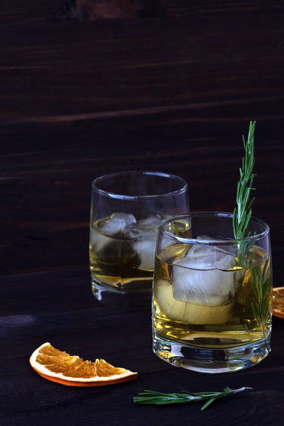 lemonade in a two glasses with pieces of ice and a sprig of rosemary on a dark background. glass of whiskey or cocktail with ice with slices of orange on a wooden table.  - Foto, imagen