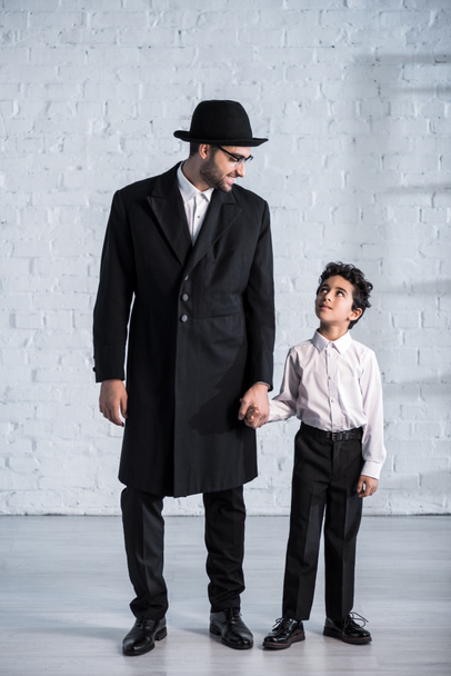 uśmiechnięty żydowski ojciec i słodki syn trzymający się za ręce i patrzący na siebie  - Zdjęcie, obraz