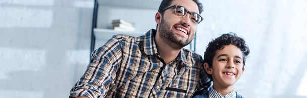 panoramic shot of smiling jewish father and son watching tv in apartment  - Photo, Image