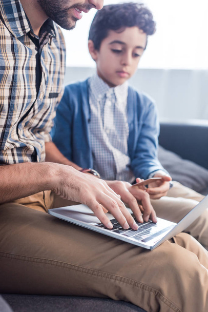 bijgesneden weergave van joodse vader met behulp van laptop en zoon met creditcard in appartement  - Foto, afbeelding