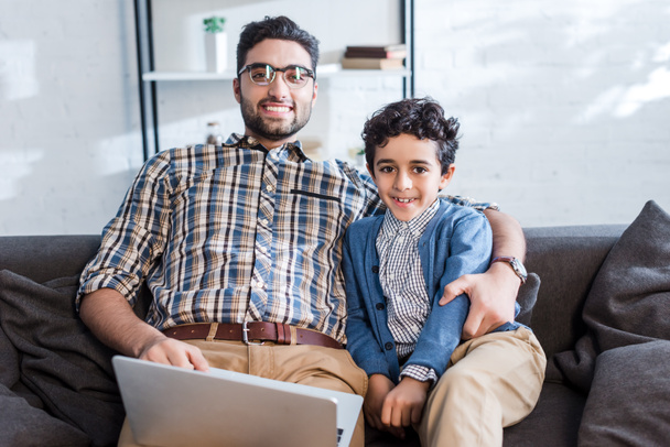 glimlachende joodse vader met laptop en zoon op zoek naar camera in appartement  - Foto, afbeelding