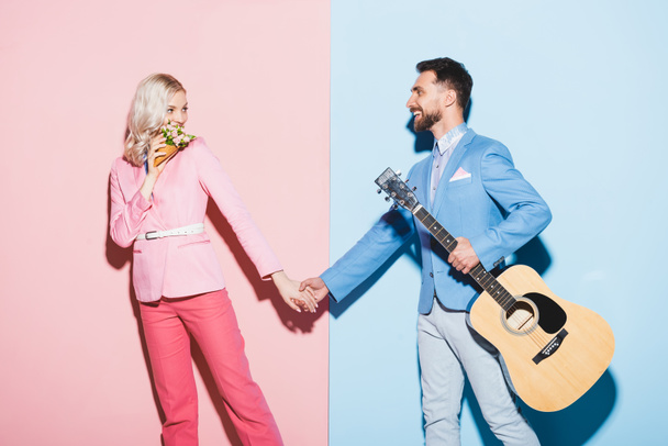 smiling woman with bouquet and handsome man with acoustic guitar holding hands on pink and blue background  - Foto, afbeelding