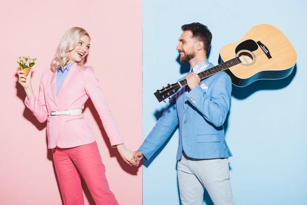smiling woman with bouquet and handsome man with acoustic guitar holding hands on pink and blue background  - Photo, Image