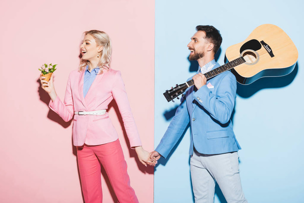 shocked woman with bouquet and handsome man with acoustic guitar holding hands on pink and blue background  - Fotó, kép