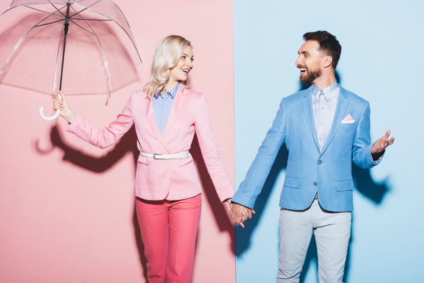 smiling woman with umbrella and handsome man holding hands on pink and blue background  - Foto, imagen