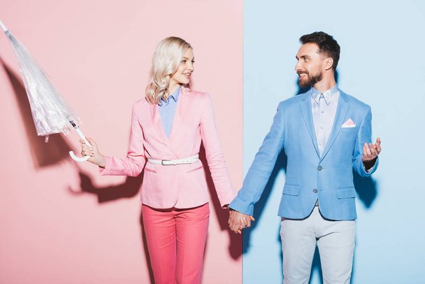 smiling woman with umbrella and handsome man holding hands on pink and blue background  - Fotografie, Obrázek
