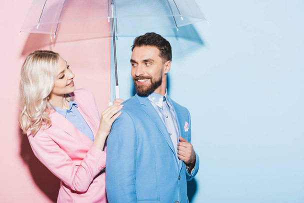 χαμογελαστή γυναίκα με ομπρέλα κοιτάζοντας όμορφο άνδρα σε ροζ και μπλε φόντο  - Φωτογραφία, εικόνα