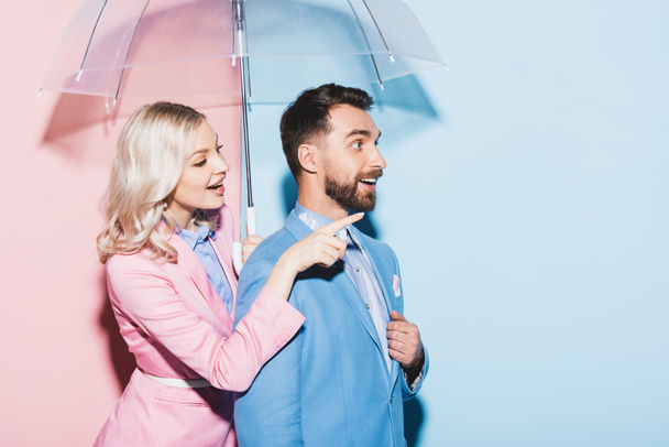 χαμογελαστή γυναίκα με ομπρέλα που δείχνει με το δάχτυλο και σοκαρισμένος άνθρωπος σε ροζ και μπλε φόντο - Φωτογραφία, εικόνα