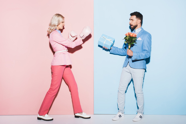 Wütende Frau mit Boxhandschuhen kämpft mit verängstigtem Mann mit Geschenk und Strauß auf rosa und blauem Hintergrund - Foto, Bild