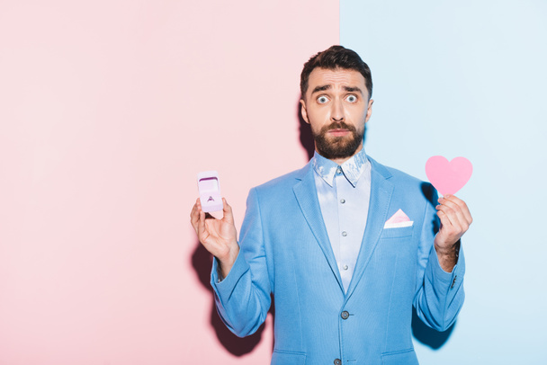 scared man holding heart-shaped card and engagement ring on blue and pink background  - 写真・画像