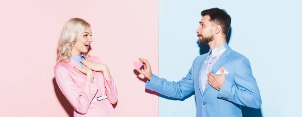 colpo panoramico di bell'uomo che dà carta a forma di cuore alla donna scioccata su sfondo rosa e blu
  - Foto, immagini