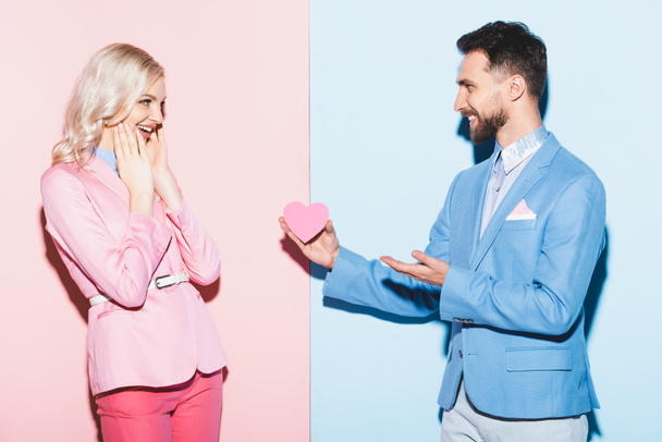 handsome man giving heart-shaped card to shocked woman on pink and blue background  - Photo, Image