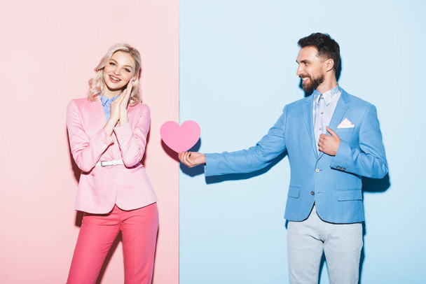 knappe man geeft hartvormige kaart aan lachende vrouw op roze en blauwe achtergrond  - Foto, afbeelding