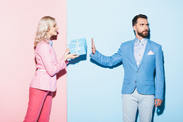 smiling woman giving gift to offended man on pink and blue background  - Photo, Image