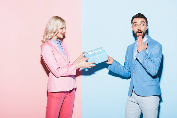 χαμογελαστή γυναίκα δίνει δώρο στον σοκαρισμένο άντρα σε ροζ και μπλε φόντο  - Φωτογραφία, εικόνα