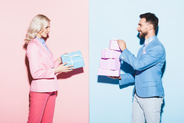 side view of attractive woman and smiling man holding gifts on pink and blue background  - 写真・画像
