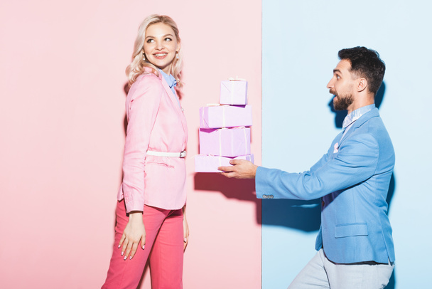 όμορφος άντρας δίνει δώρα σε χαμογελαστή γυναίκα σε ροζ και μπλε φόντο  - Φωτογραφία, εικόνα