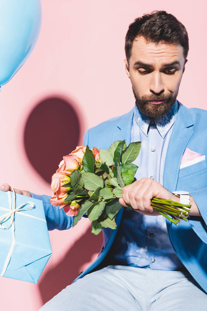 knappe en nerveuze man op zoek naar polshorloge en het houden van ballon, cadeau en op boeket blauw en roze achtergrond  - Foto, afbeelding