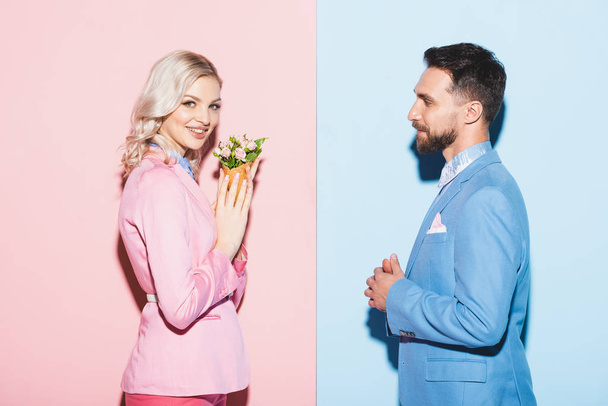 boční pohled na usmívající se žena s kyticí a pohledný muž na růžovém a modrém pozadí  - Fotografie, Obrázek