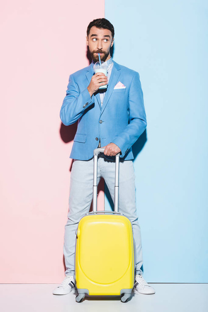 man drinking cocktail and holding travel bag on pink and blue background  - Foto, afbeelding