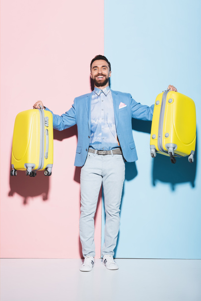 χαμογελαστός άνδρας κρατώντας τσάντες ταξιδιού σε ροζ και μπλε φόντο  - Φωτογραφία, εικόνα