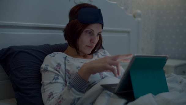Young female brunette adult with sleeping mask on head surfing online in tablet computer in bed. Woman taking away gadget and going to sleep.  - Imágenes, Vídeo