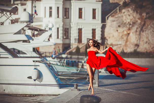 Model girl in a long red flowing dress walks along the waterfront with yachts - Photo, Image