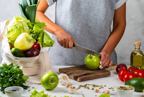 Kadın yeşil bir elma keser ve masada taze sebzelerle dolu bir pamuk torbayla sağlıklı bir diyet yemeği pişirir.. - Fotoğraf, Görsel
