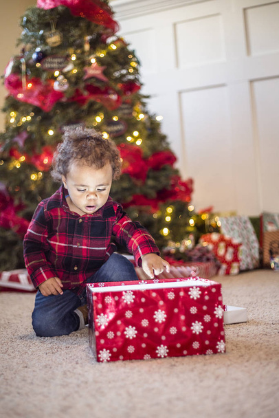 Aranyos kis sokszínű fiú kinyitja a varázslatos karácsonyi ajándékot egy karácsonyfa előtt. Izgatott arckifejezés a gyermek arcán, ahogy belenéz a dobozba. - Fotó, kép
