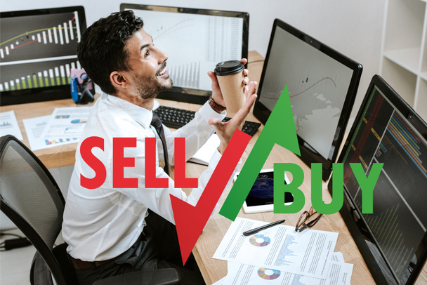 happy bi-racial trader holding paper cup and sitting near computers and sell, buy letters - Photo, Image