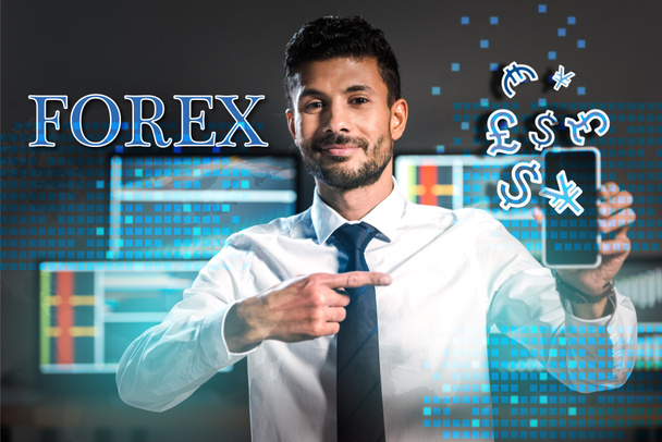 smiling bi-racial trader pointing with finger at smartphone near forex letters and money signs  - Photo, Image