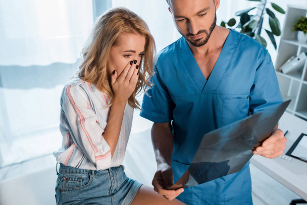 ortopedista guapo y mujer molesta cubriendo la cara mientras mira rayos X
  - Foto, Imagen