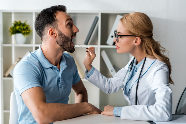 side view of attractive doctor in glasses examining man with medical spatula  - Фото, зображення