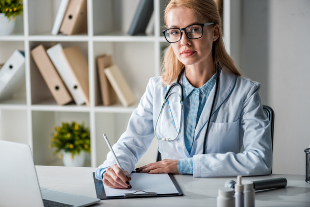 attraente medico in occhiali scrittura diagnosi vicino laptop
  - Foto, immagini