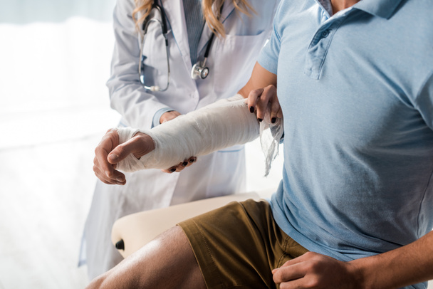 cropped view of orthopedist putting bandage on injured hand of man  - Fotografie, Obrázek