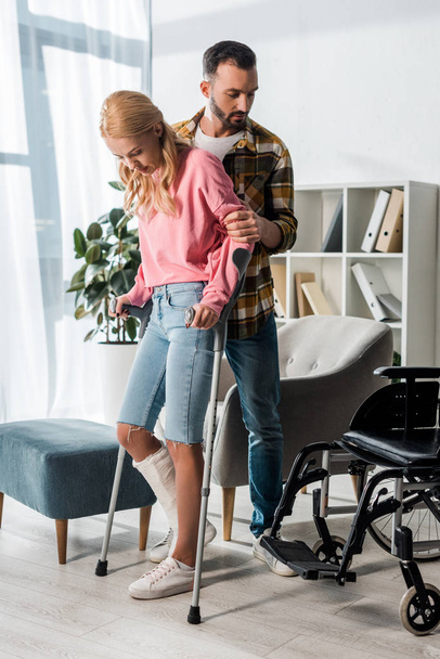 injured woman holding crutches while standing with man near wheelchair at home - Photo, Image