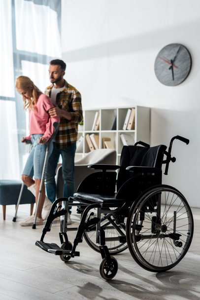 selective focus of wheelchair near woman holding crutches while standing with man - Photo, Image